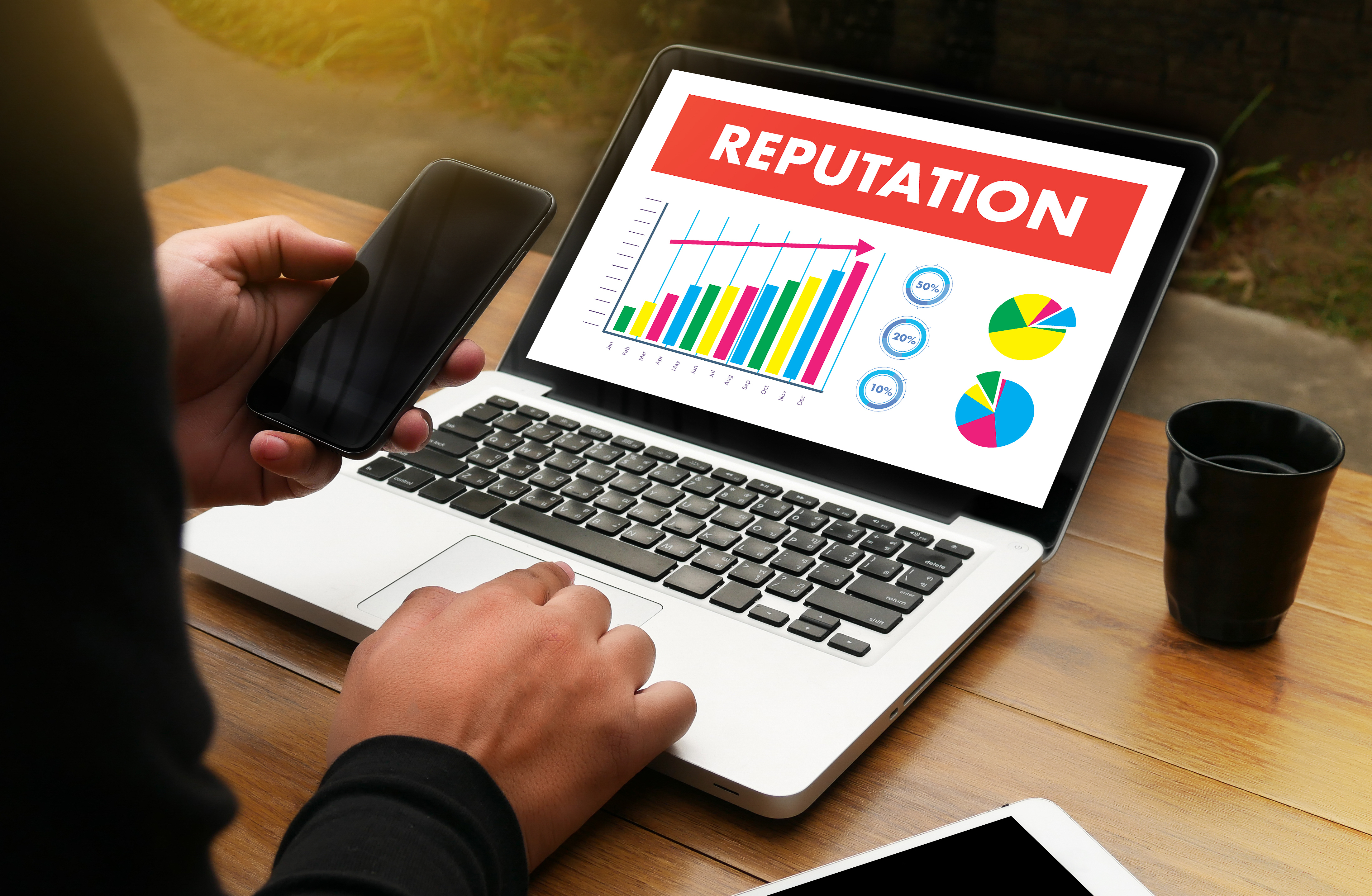 A person sitting at a laptop. On the laptop screen there is a barchart, with a red arrow pointing in an upwards direction, with the word 'reputation' above, indicating the person's reputation is increasing